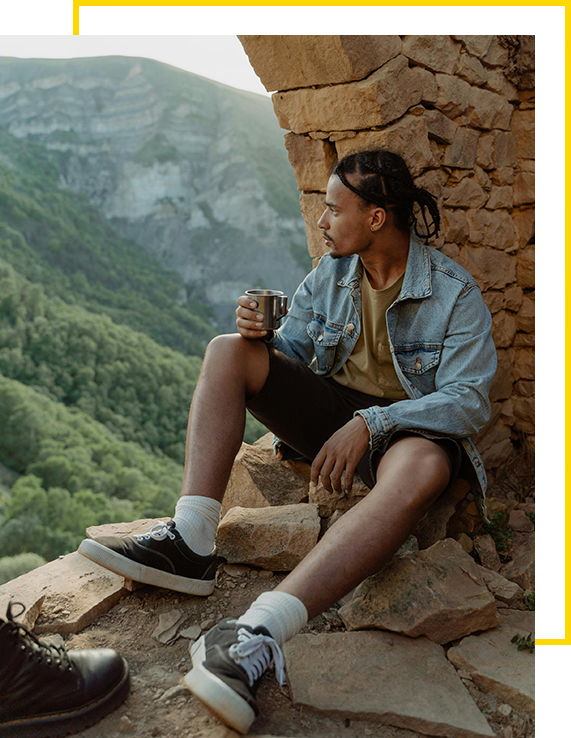 Person sitting on a ledge holding a cup, overlooking a green mountainous landscape.